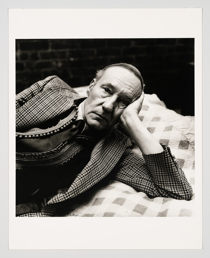 An older man lying on a patterned blanket, resting his head on his hand and looking thoughtful.