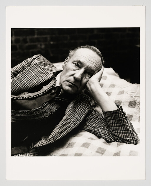 An older man lies on a patterned blanket, resting his head on his hand and looking thoughtful.