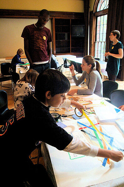A child adding tape to an artwork on a table, with other people in the background.