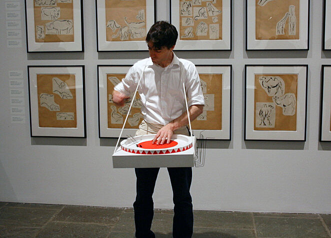 A man in a white shirt stands in front of a wall of prints, with some sort of contraption strapped to their body.