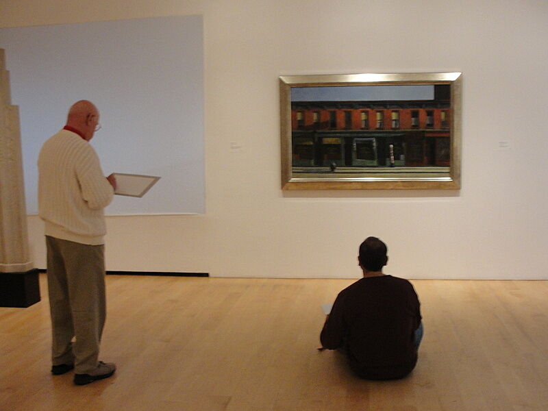 A man stands and a student sits on the gallery floor in front of a painting.