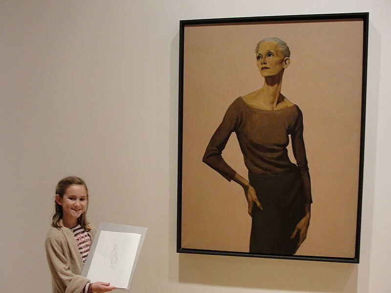 A girls shows off her artwork in front of a painting of a woman.