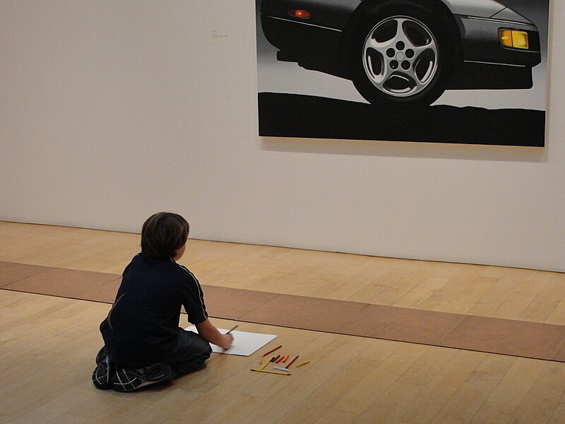 A child sits and draws in front of an artwork depicting a car.