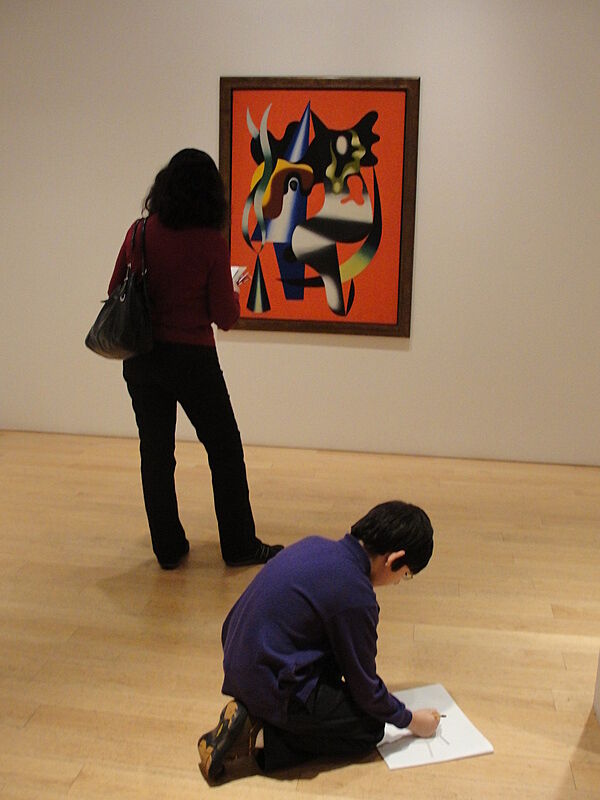 A boy sits on the floor to draw a picture, while a woman stands in front of a modern painting.