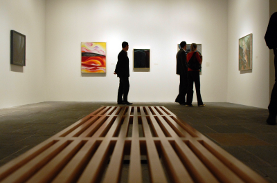 The view down a bench in the gallery towards a wall with paintings, and few people milling about.