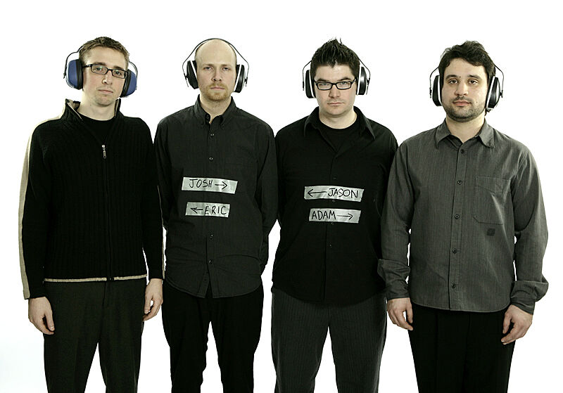 Four men stand without expression in a row, all wearing hearing protection ear muffs.