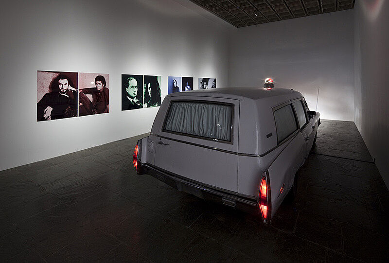 A white hearse parked in the gallery with it's taillights on and photographs on of people on the wall behind.