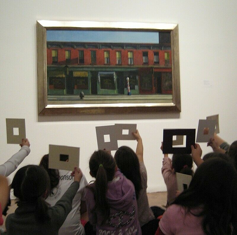 A group of children hold up pieces of paper with rectangular holes cut into them in front of a painting of a building.