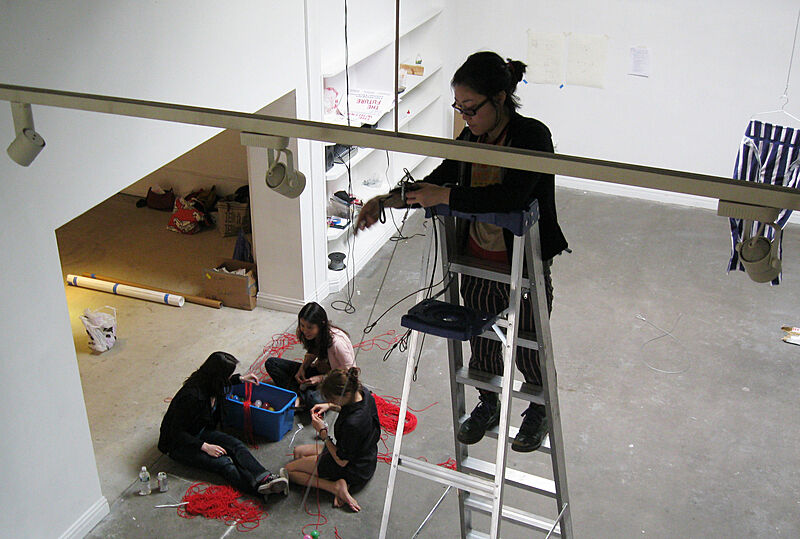The artist stands on a ladder while kids sit on the ground and work with string.