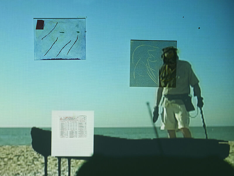 A video projection of a man with a metal detector on the beach, on top of other materials and documents.