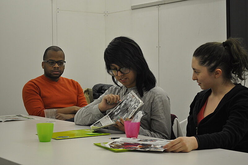 A teen shows her newspaper art project to the artist at a table.