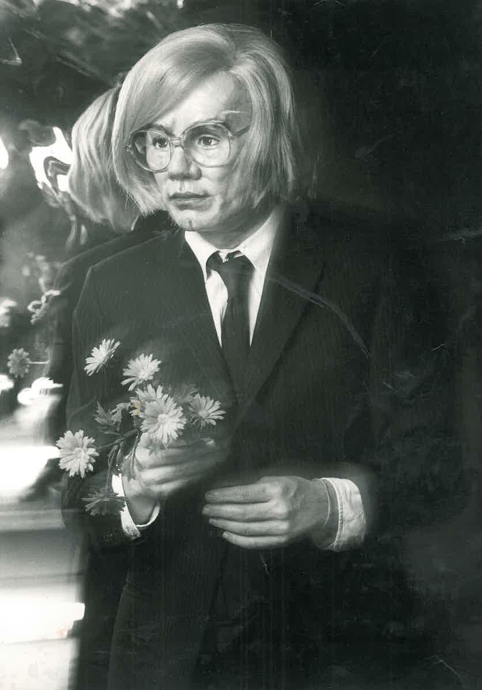 Person with blond hair and round glasses holding daisies, wearing a suit and tie.