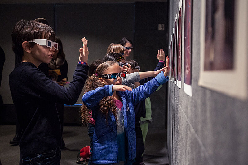 Children wearing 3D glasses attempt to interact with art on a wall.