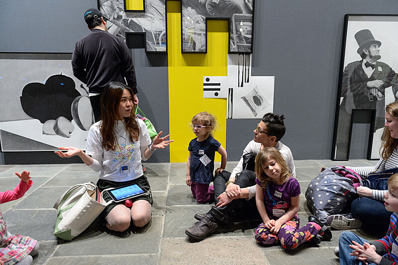 Kids and adults sit on the floor and listen to a museum staff member in a gallery.