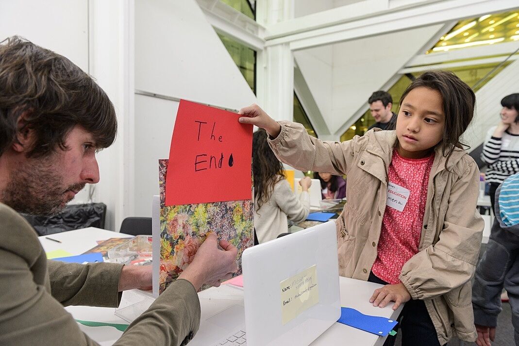 A child holds up a piece of paper which reads "The End" in front of a laptop.