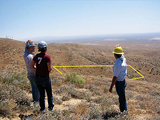 Three people wearing hard hats stand on a dry hillside, looking at a marked rectangular area below.