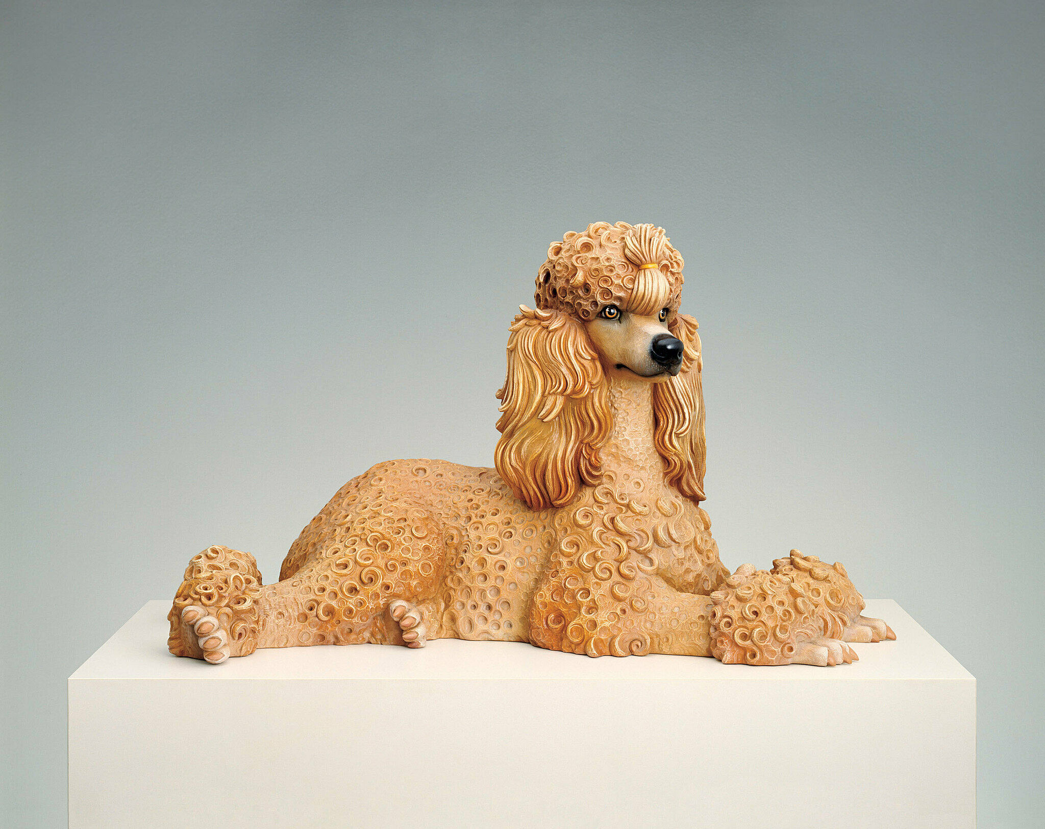 A realistic sculpture of a poodle with curly fur, lying on a white pedestal against a plain gray background.