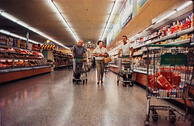 Three shoppers with carts in a vintage grocery store aisle, surrounded by products on shelves.