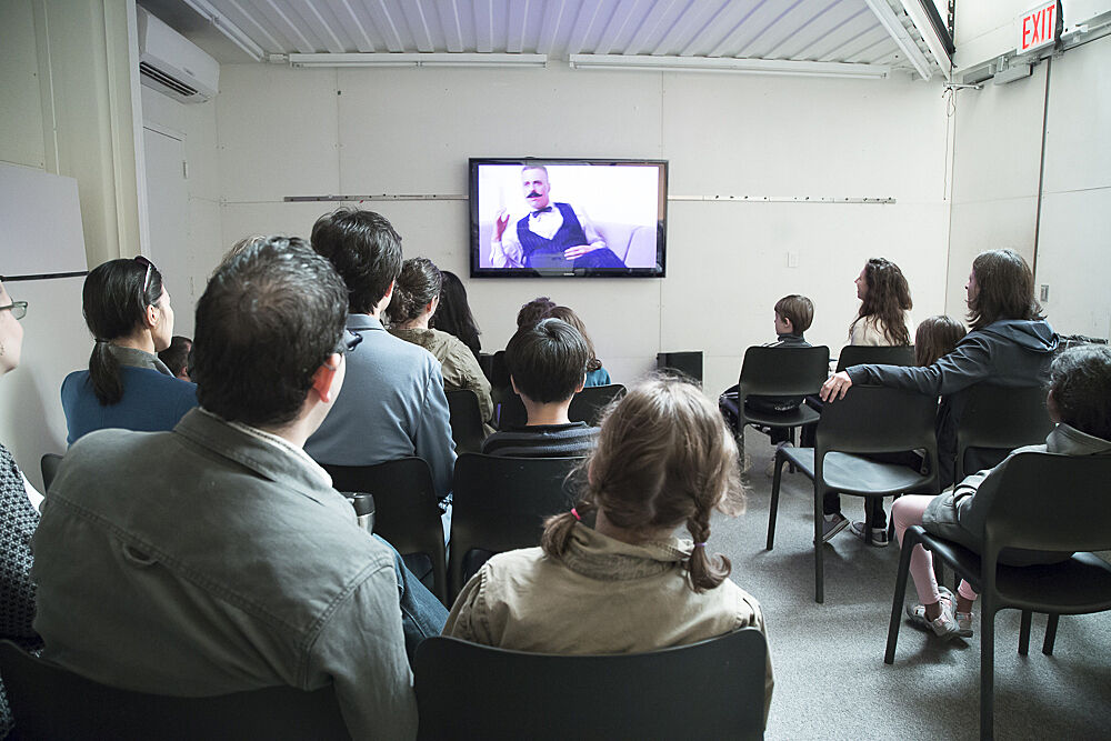 A group of visitors watch the film on a flatscreen TV at the front of a room