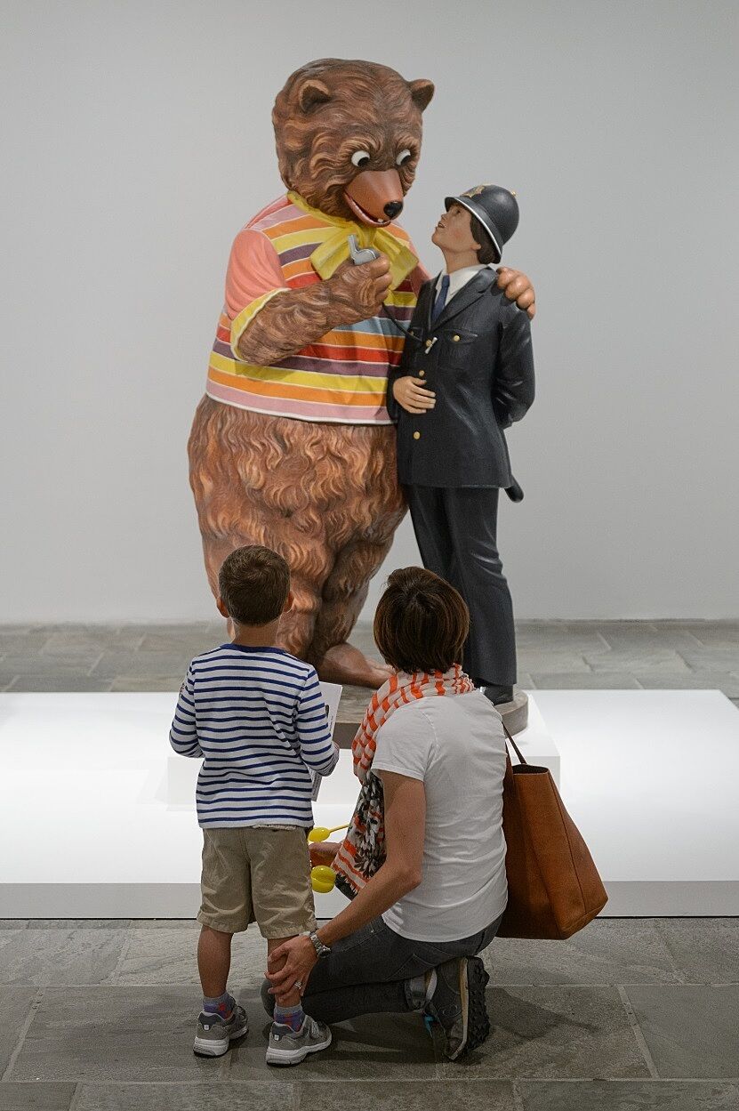 A mother and child stop in front of a bear and policewoman sculpture by Jeff Koons.