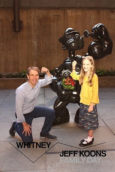 Jeff Koons and a young girl hold up their arms in front of a sculpture of Popeye.