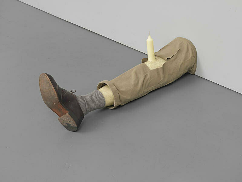 An installation of a leg sticking out of a wall with a wax candle sticking out of the knee.