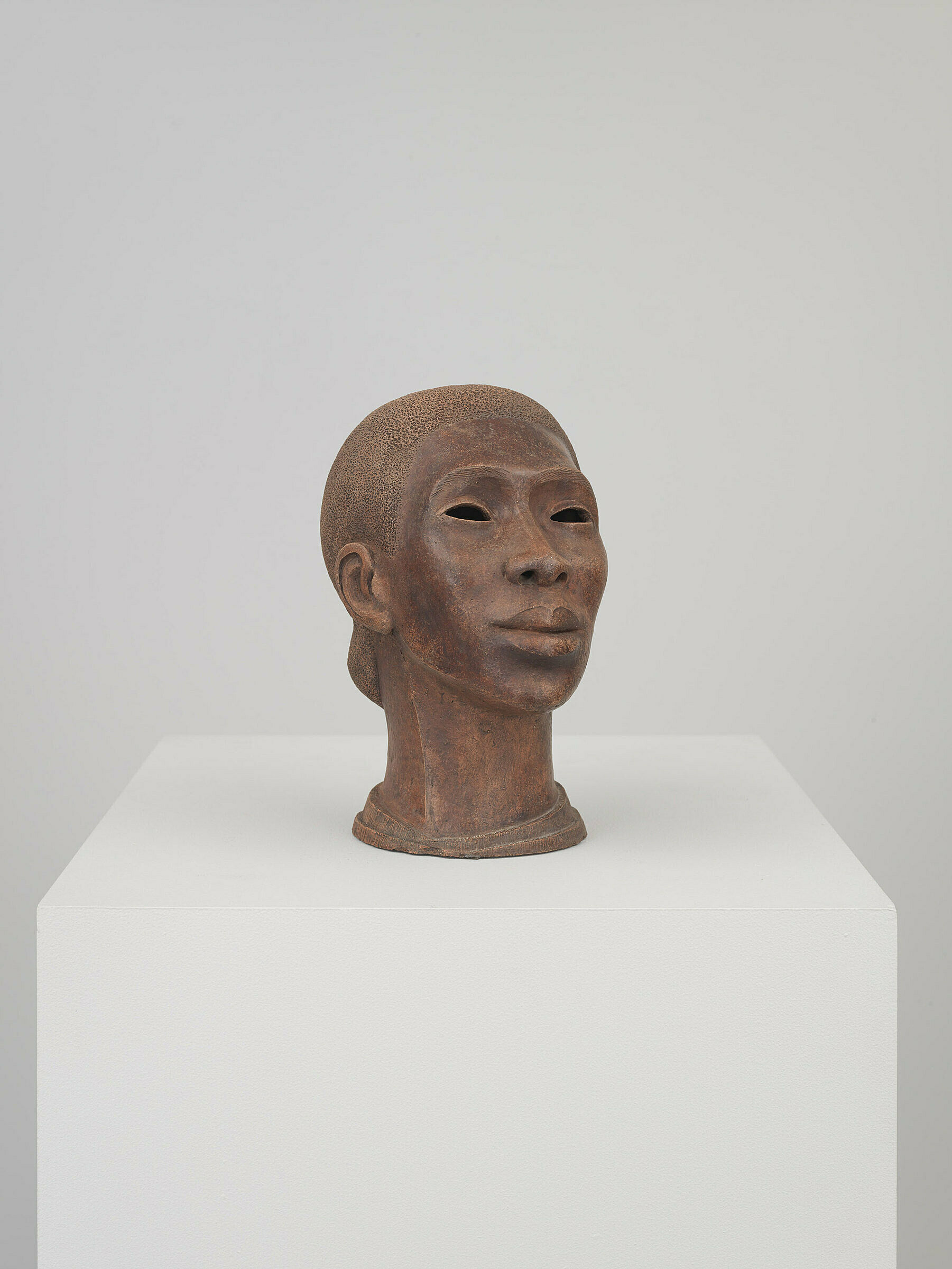 A brown terracotta sculpture of a woman's head down to the neck sits on a white plinth. The eyes of the sculpture are cut out.