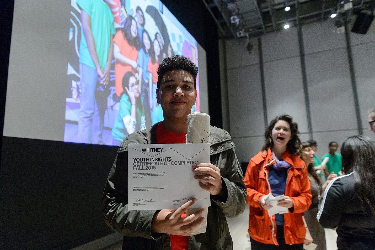 Person holding a certificate with a proud smile, in front of a projected group photo.