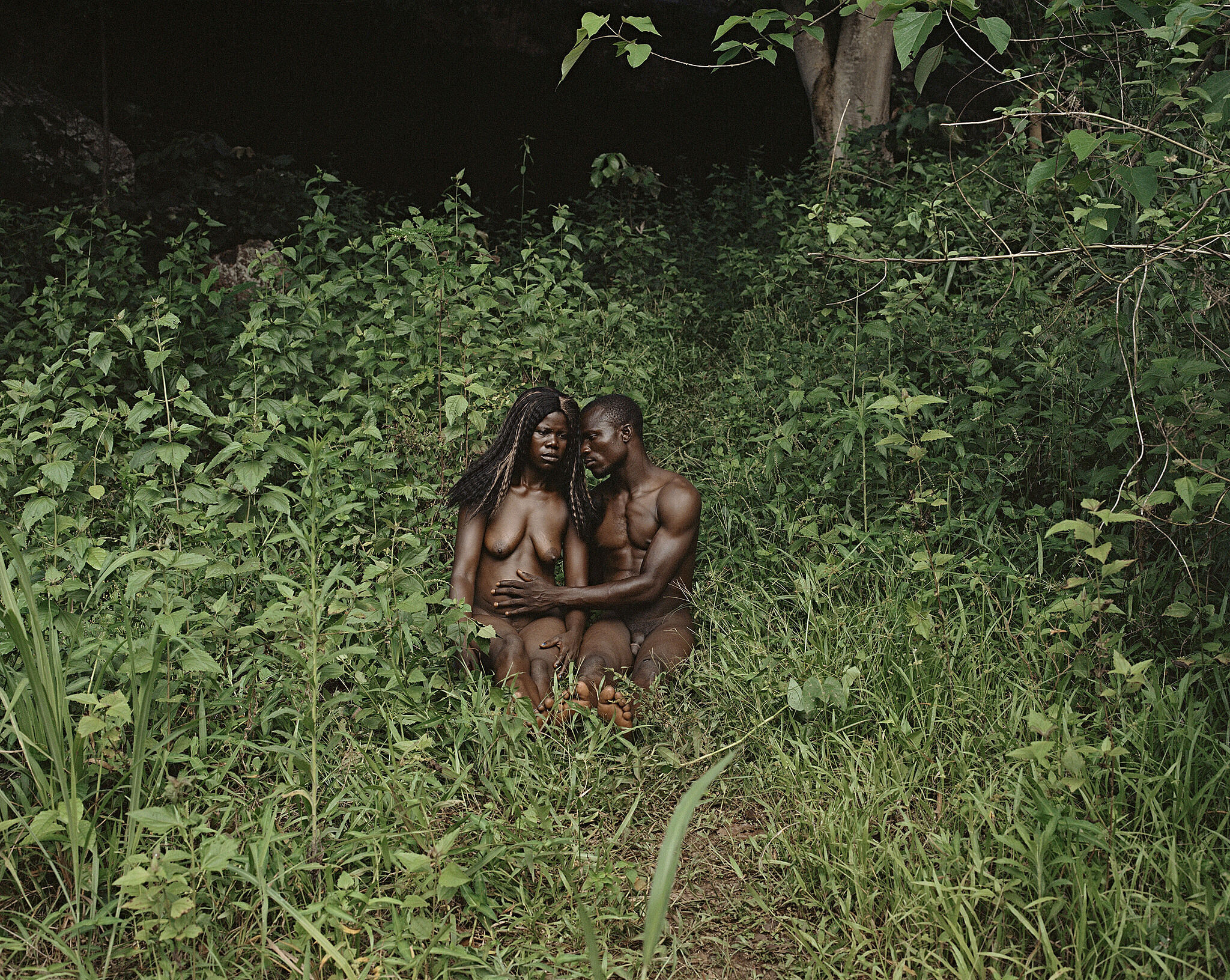 A Black couple sit in the nude in a lush green garden; the man's hand reaches across the woman's belly.