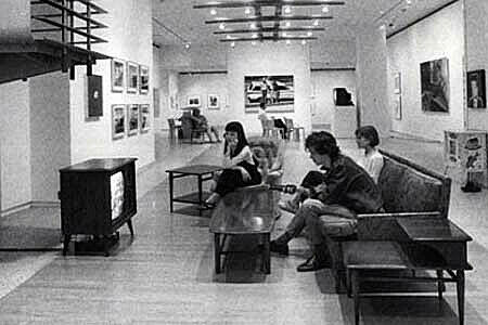 Three people sit on couches in a quiet gallery watching a small television.