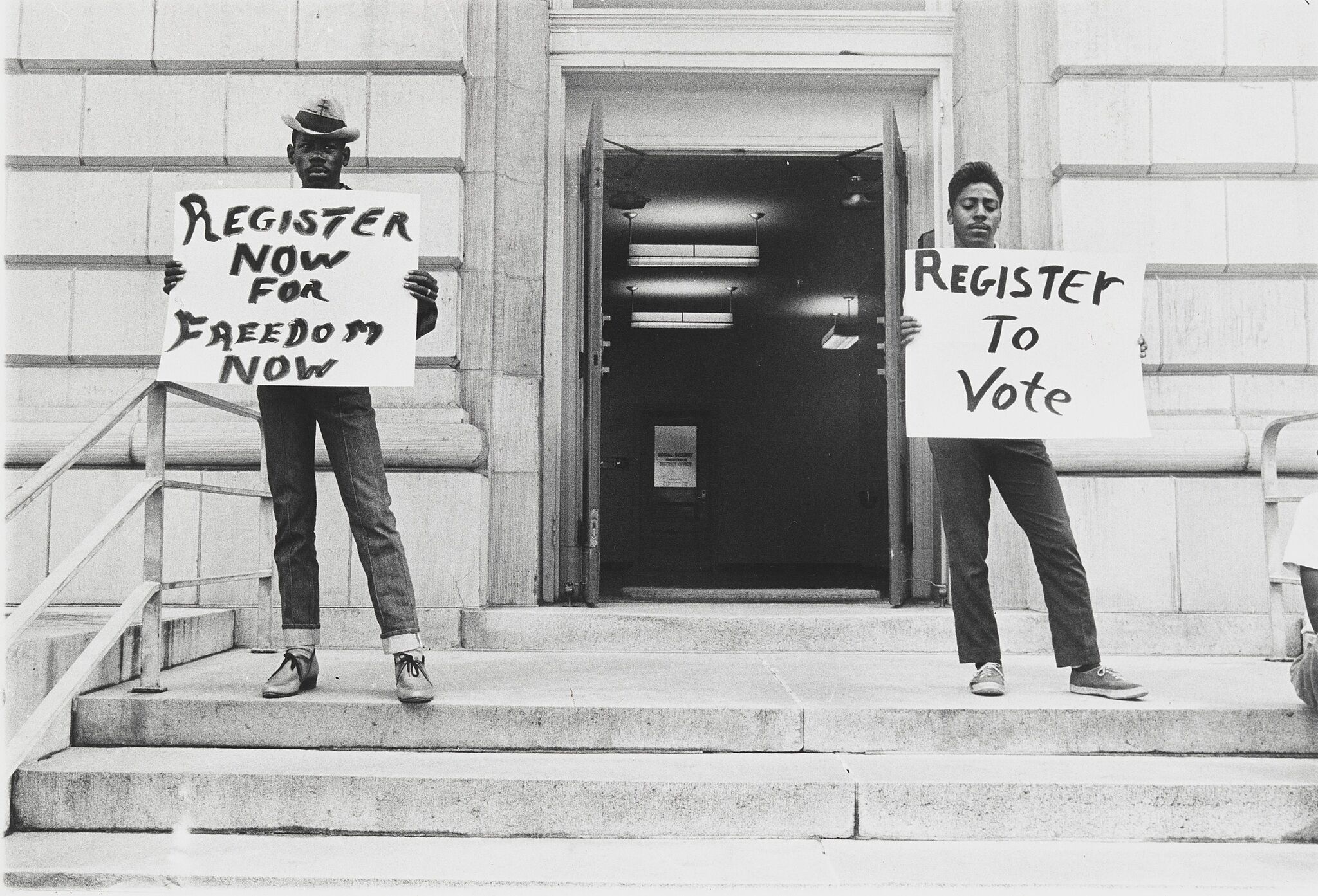 Two people holding signs that say "Register now for freedom now" and "Register to vote"