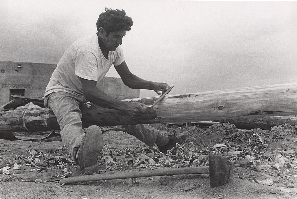 A photograph of a man shaving the bark off of a log.