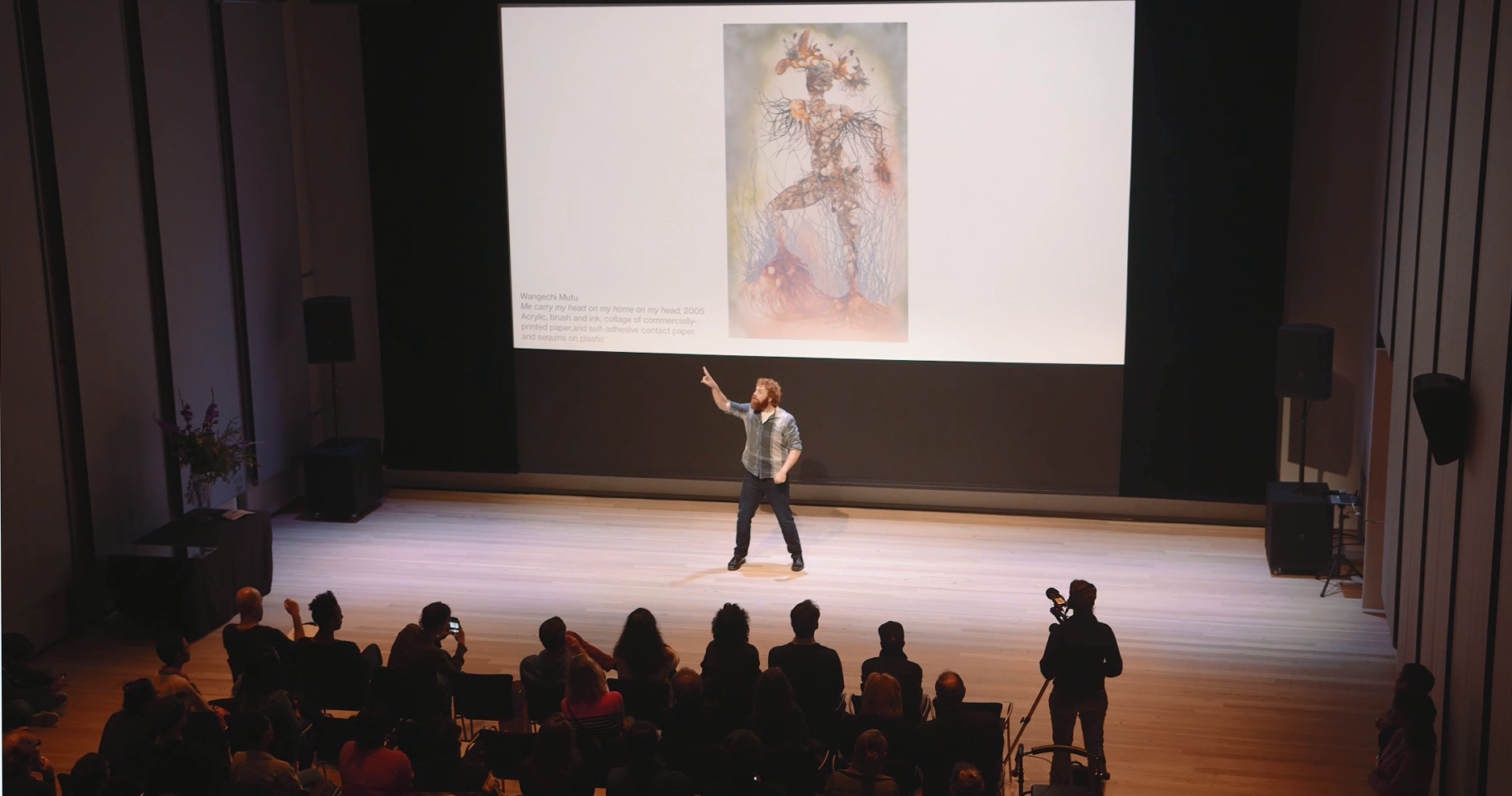 A man signs in front of a crowd with a projector screen behind him.