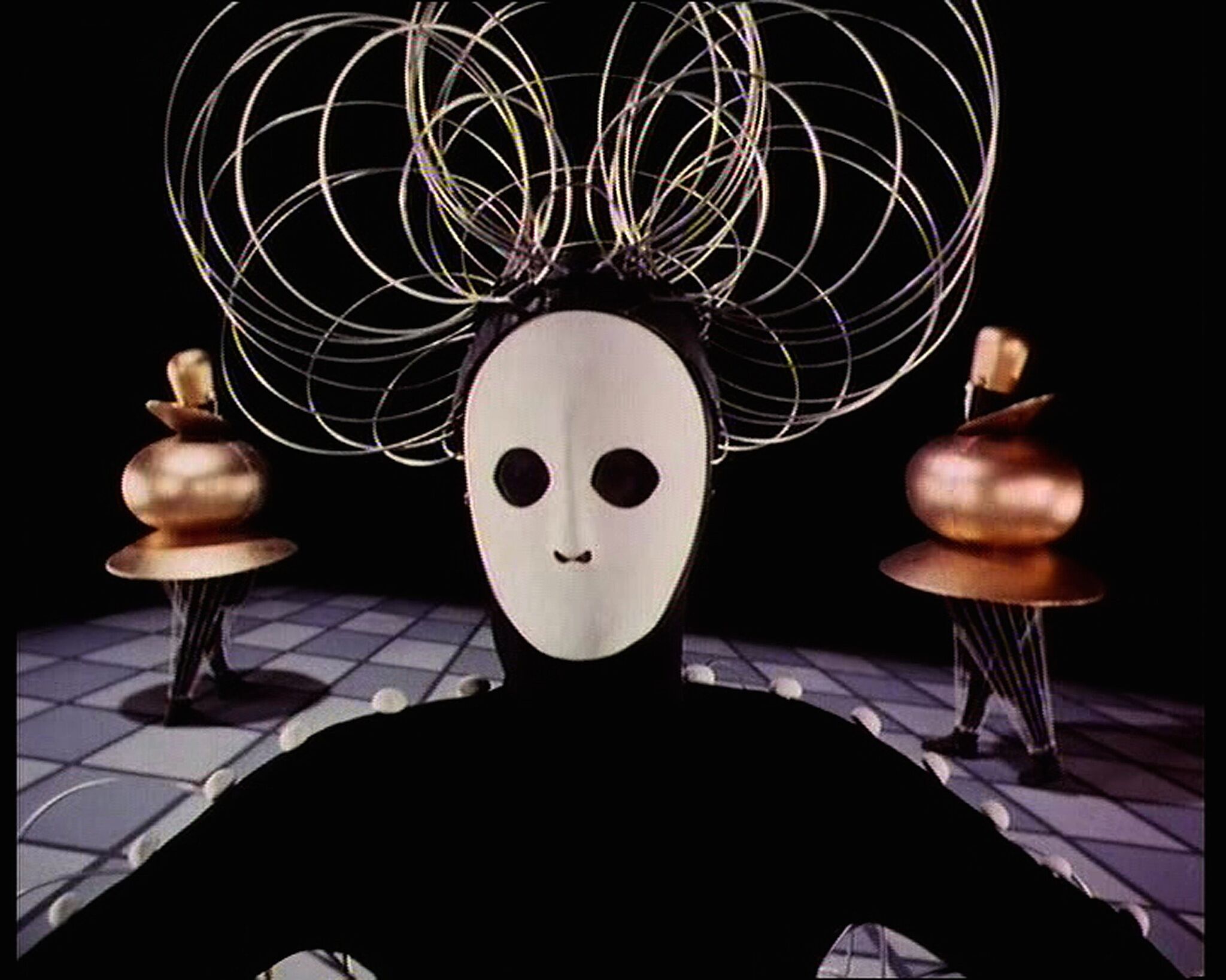 Film still of a dancer figure dressed in black and wearing a white mask