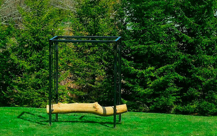 An artwork by Virginia Overton. A log is suspended by an iron structure is installed in a large field