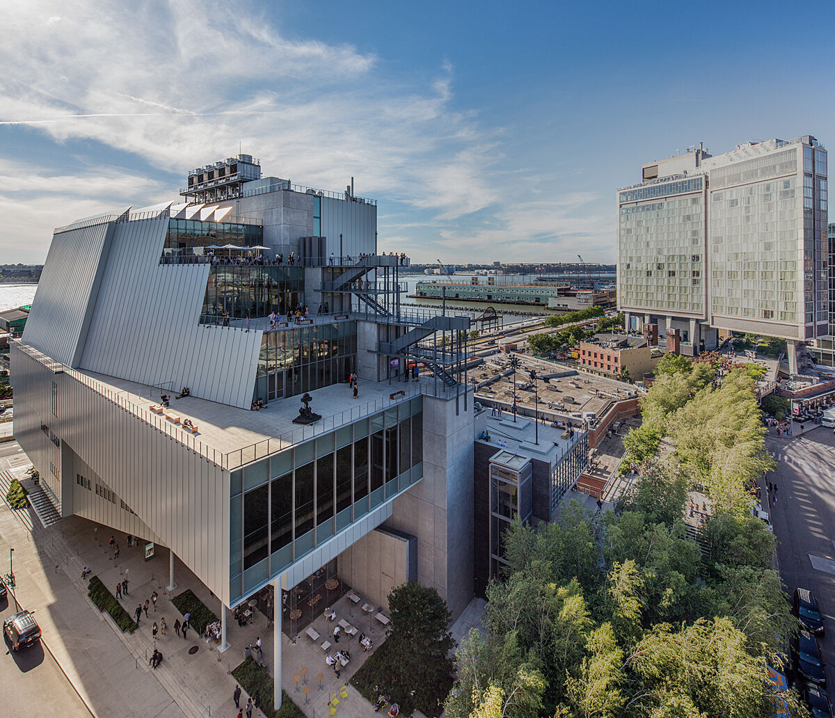 Modern building with tiered terraces and large windows, surrounded by trees and cityscape, under a clear blue sky.