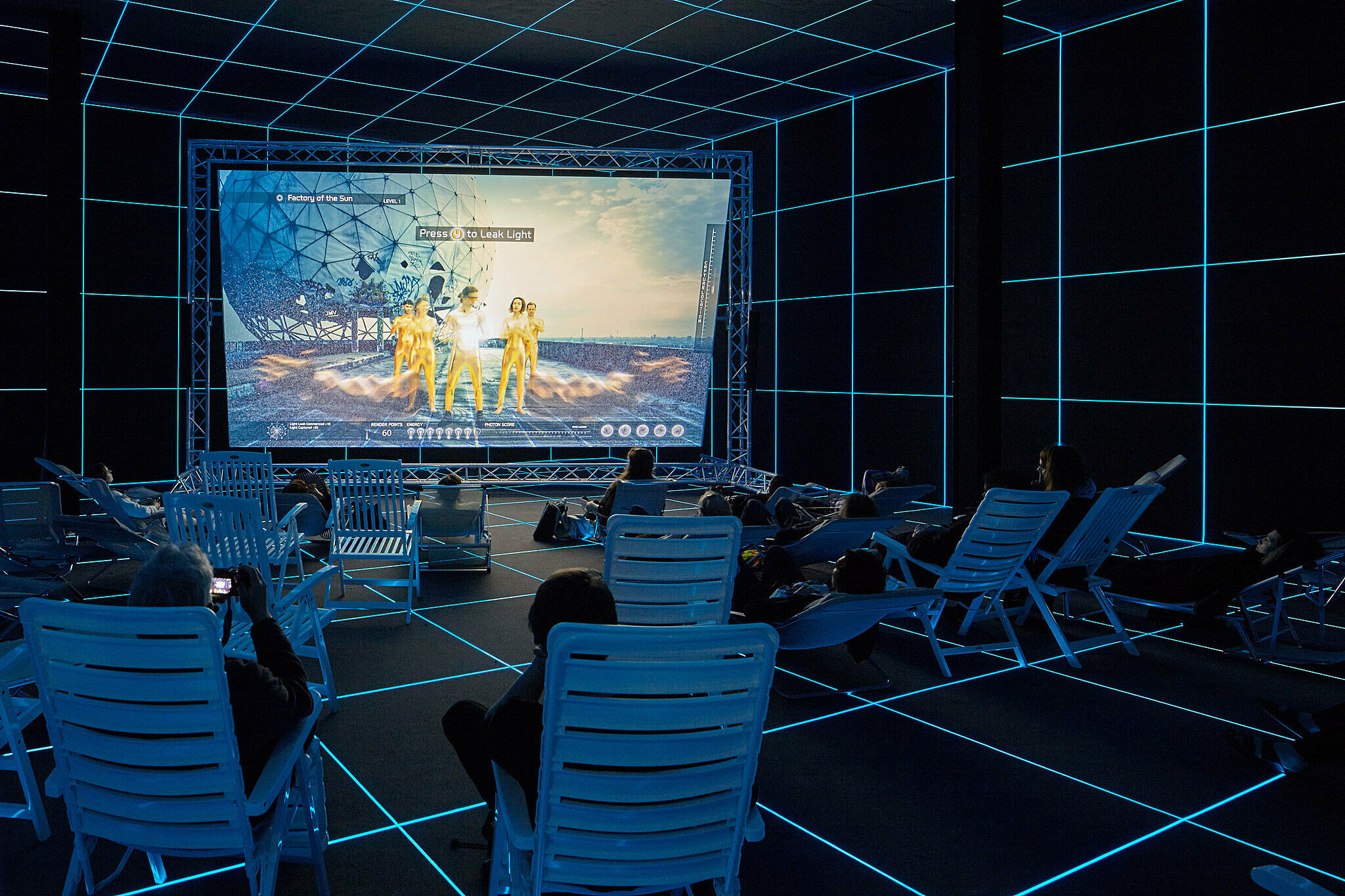 A work by Hito Steyerl. Visitors sit and recline in lawn chairs viewing a screen