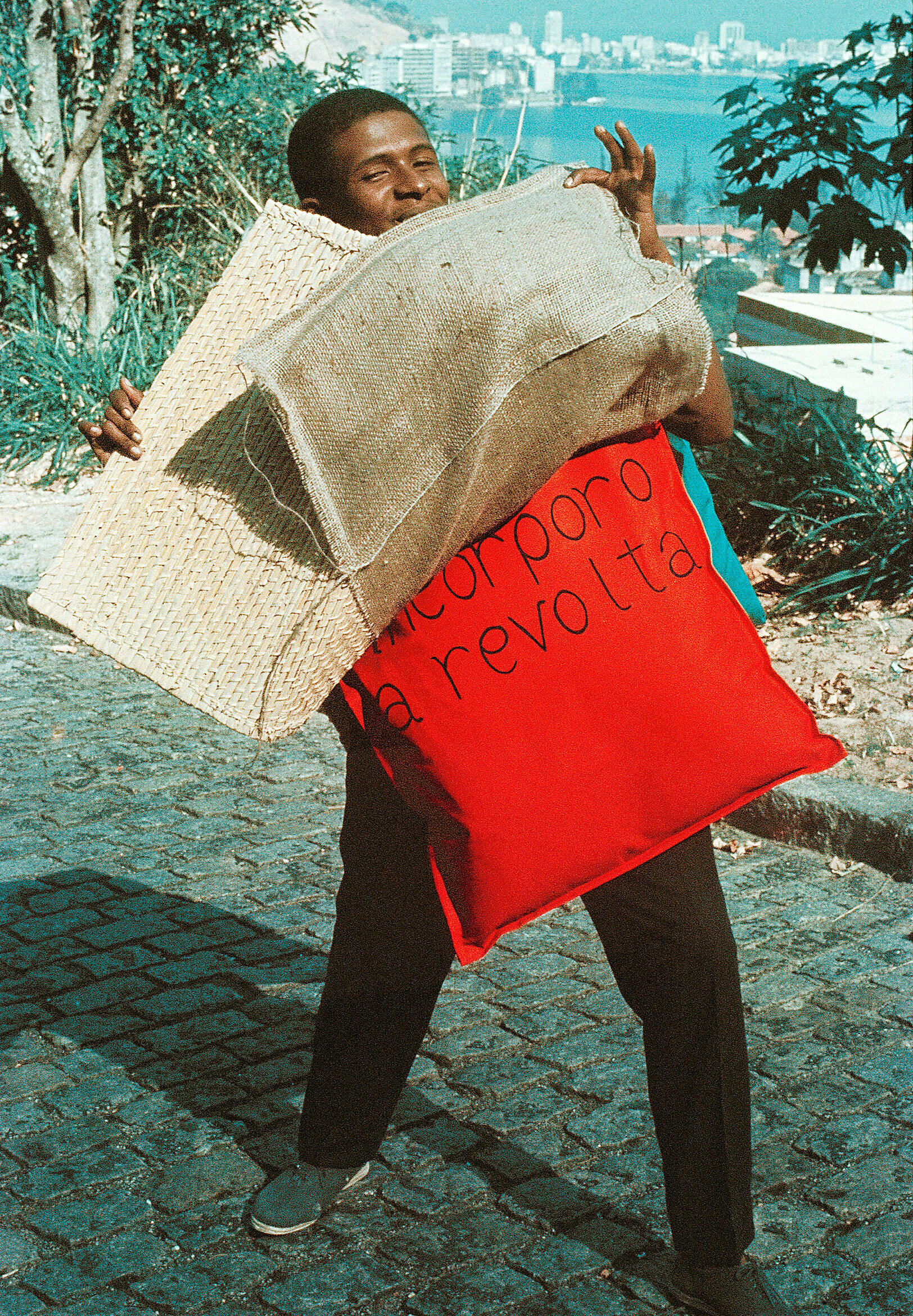 Image of man standing in street on hill overlooking body of water and holding burlap and pillows that say incorporo a revolta