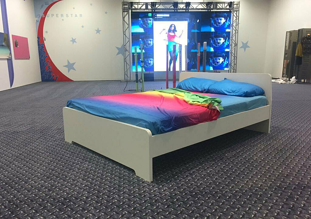 Installation view of Assymmetrical Response includes a bed with rainbow sheets and metallic floor