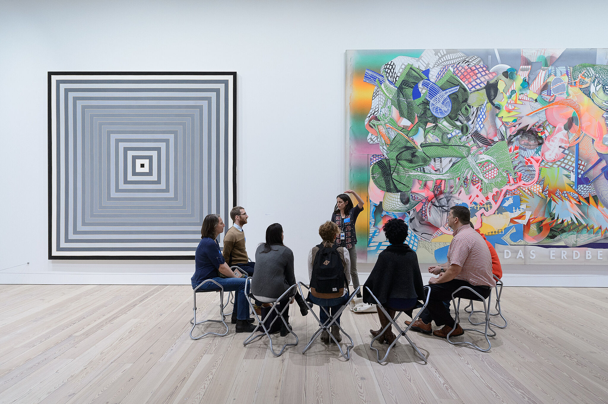 A group of people sits in front of two large artworks in a gallery, listening to a guide. One artwork is geometric, the other is abstract.