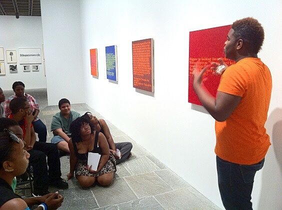 man in orange talking about paintings on wall looking at ground