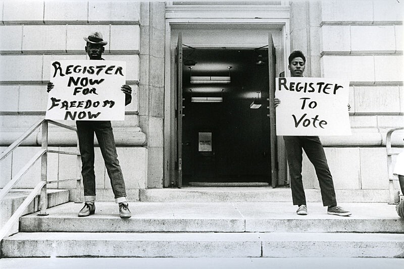 Register to vote signs held by men outside of building.