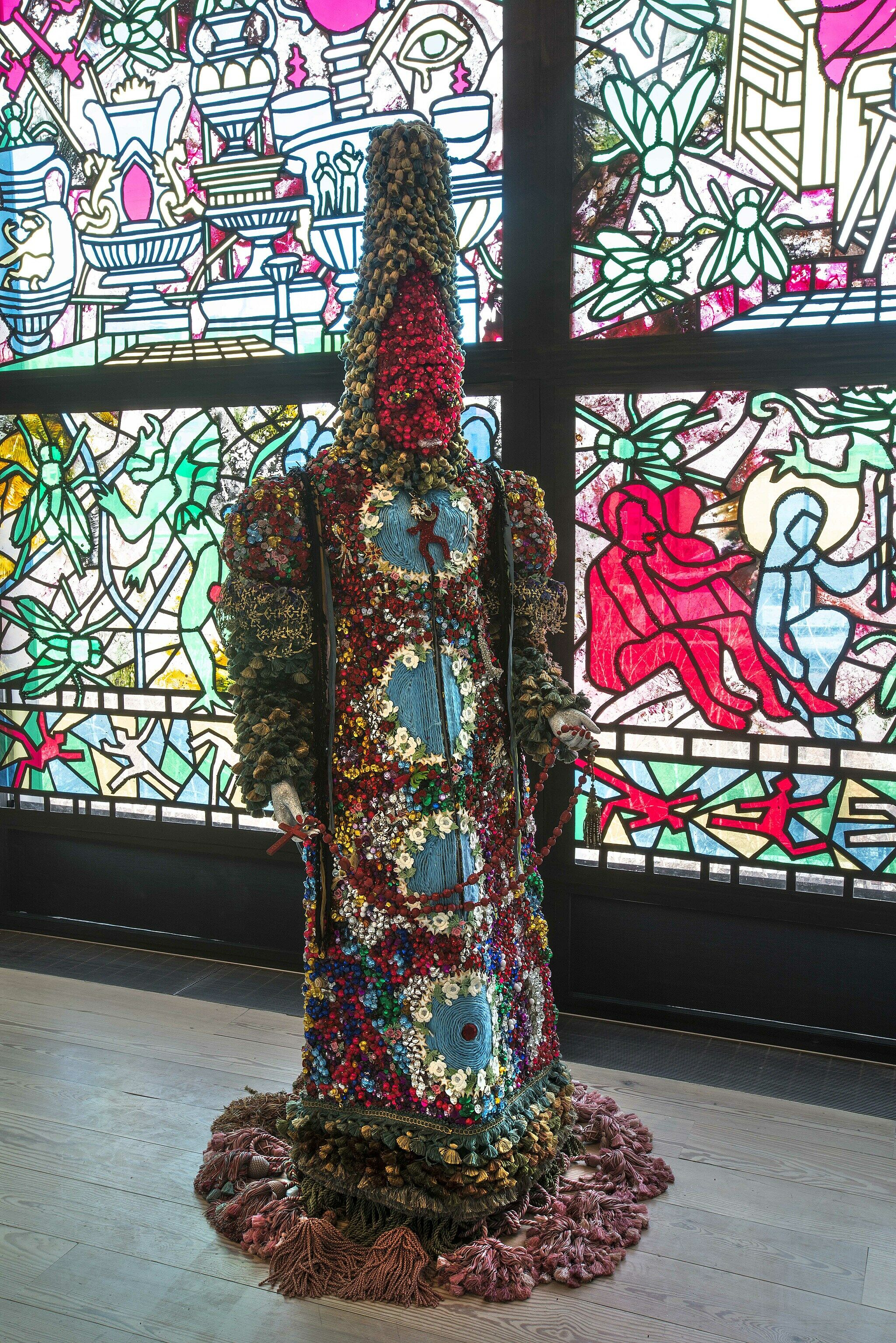 Mannequin with cone shaped head covered in beads and cloth in front of a colorful window