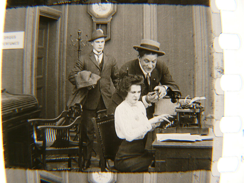 Two men look over the shoulder of a woman on a typewriter.