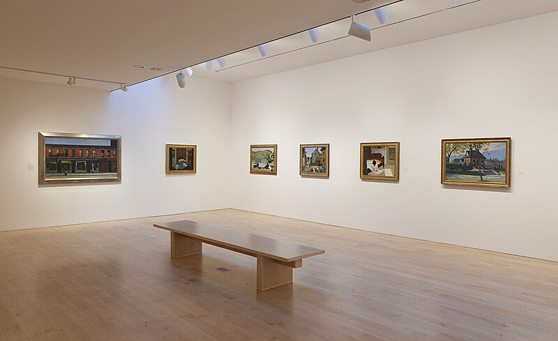 A gallery with paintings on white walls and a bench in the middle.