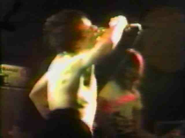 A video still of a man singing into a microphone without a shirt on.