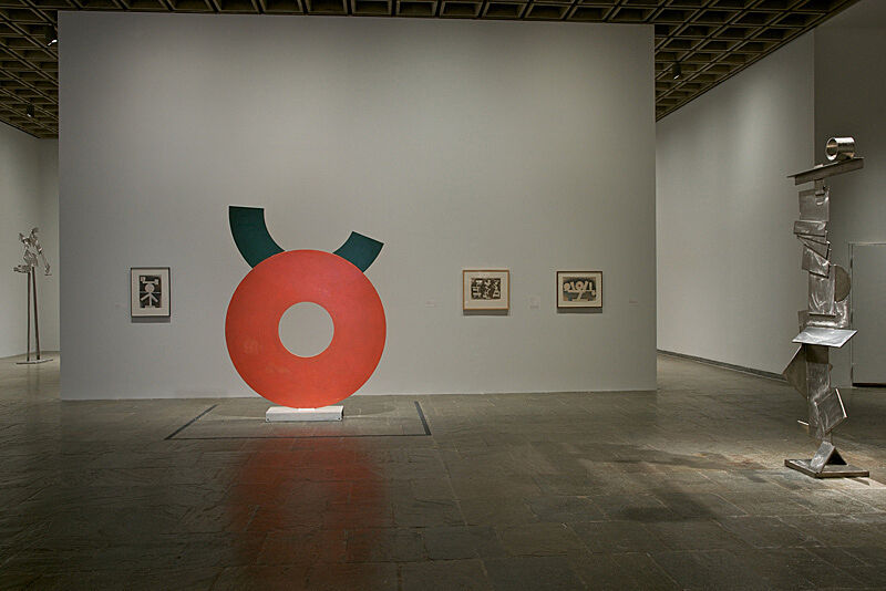 An orange and green circular sculpture and a silver sculpture stand in a gallery with three painting on the wall.