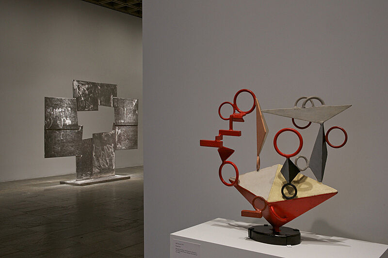 A small sculpture sits on a table while a large cube sculpture stands in the background of a gallery.