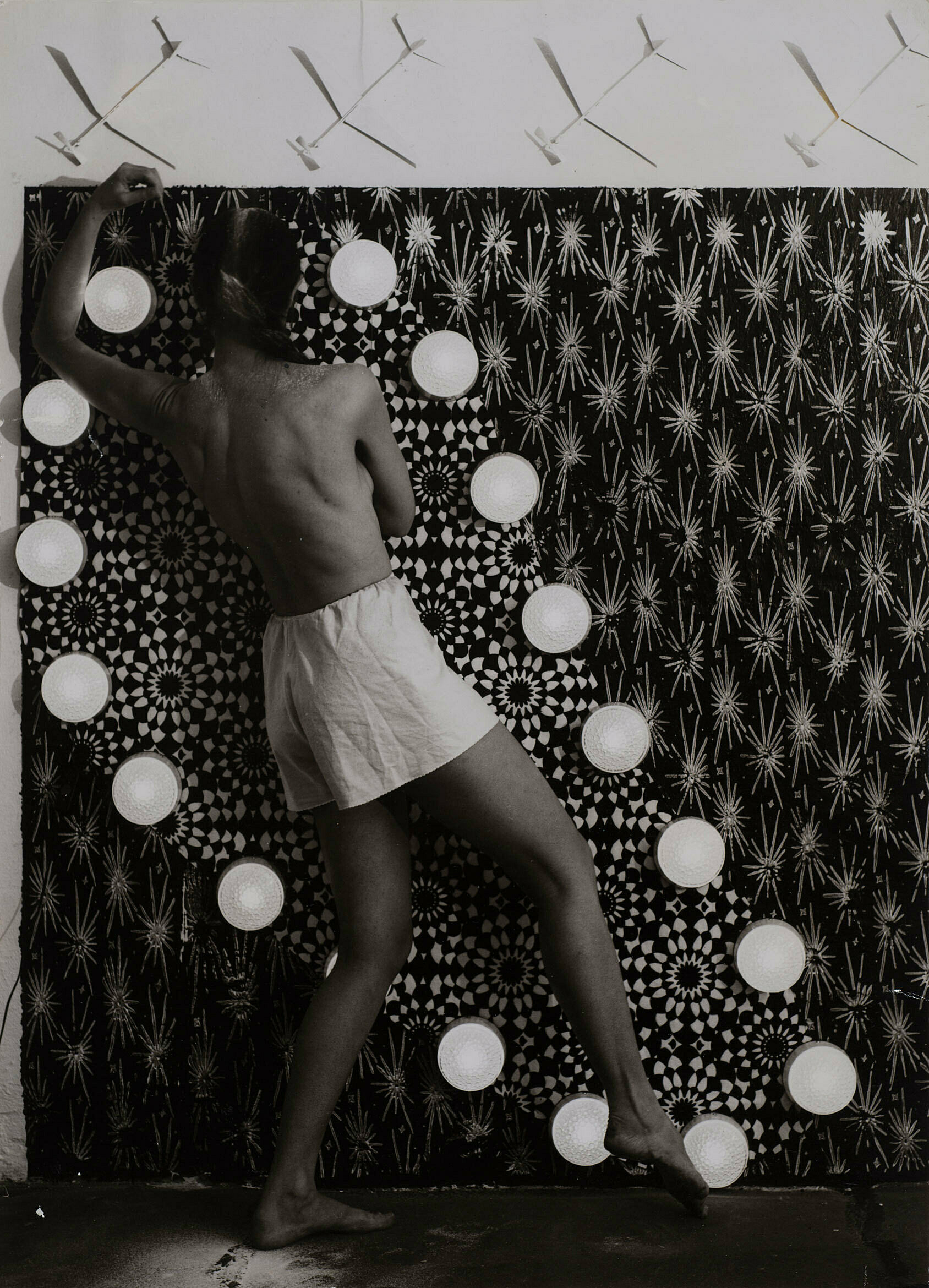 A topless woman stands with her back to the camera in front of a curtain.