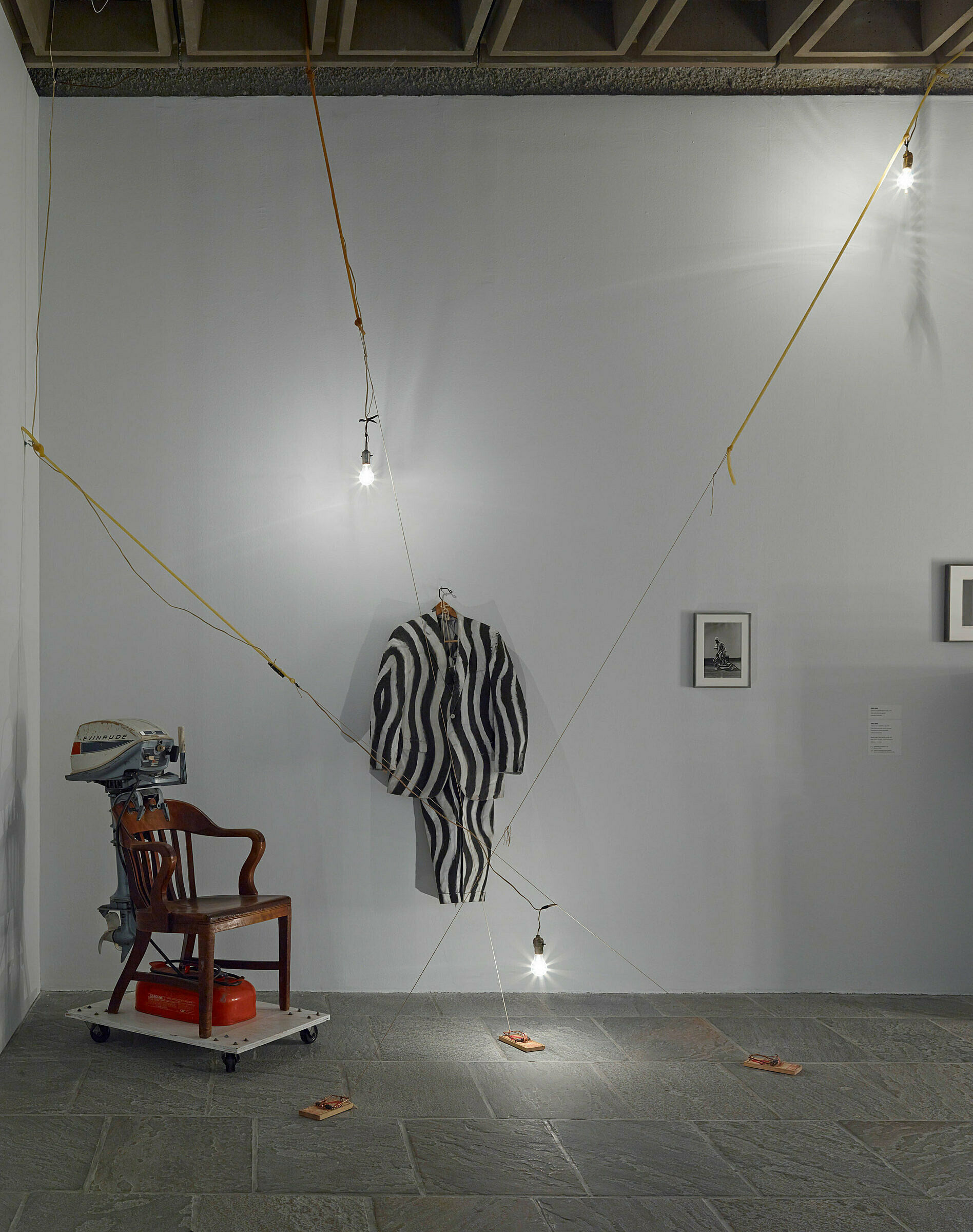 Art installation in a gallery with a chair in the corner and striped outfit on the wall.